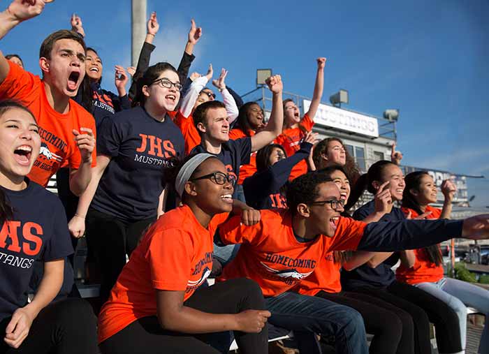 High school students cheer in the stands at a football game in matching custom t-shirts.